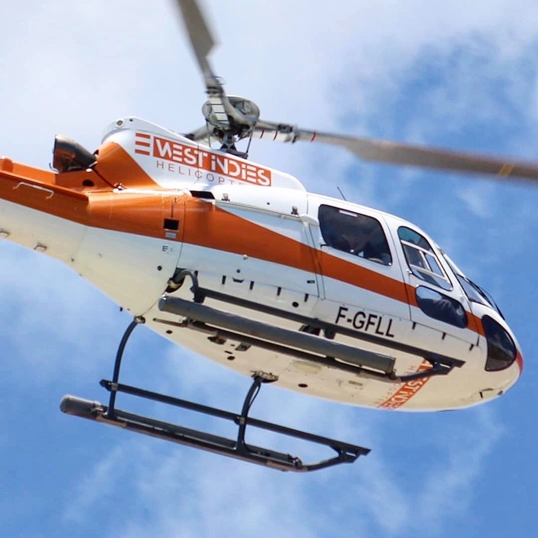 Hélicoptère blanc et orange de West Indies Helicopters en vol, vu depuis le sol